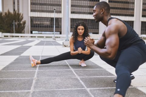 Young woman with curly hair and muscular male friend wearing sports attire, squatting and doing leg stretches, warming up together before beginning workout on checkered floor of building terrace