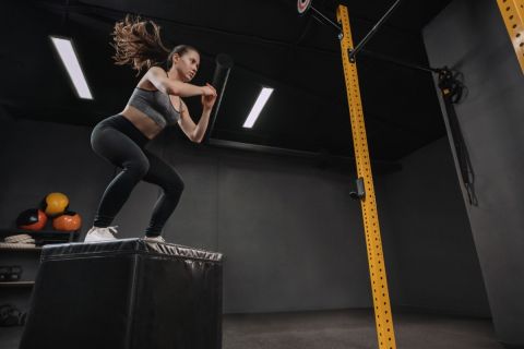 Woman doing box jump exercise as part of her gym training. Female athlete doing squats and jumping onto the box in dark workout gym. Copy space