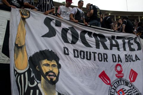 Supporters of Corinthians stand next to a huge banner in homage of the late footballer Socrates, during the Brazilian Championship final date match against Palmeiras, at the Pacaembu stadium on December 04, 2011 in Sao Paulo, Brazil. Socrates -former captain of the national team and player of Corinthians- died earlier today at the age of 57 from an intestinal infection. AFP PHOTO / Yasuyoshi CHIBA

