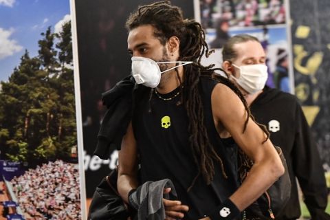 Germany's tennis player Dustin Brown wears a face mask as he walks by a poster at a pro-tennis tournament at a local base tennis academy in Hoehr-Grenzhausen, Western Germany, Friday, May 1, 2020. The professional tennis exhibition in the small village in the Westerwald is a rare exception to the global shutdown of sports during the coronavirus pandemic. Matches are played without line judges and without spectators, and broadcasted by remote cameras worldwide. (AP Photo/Martin Meissner)